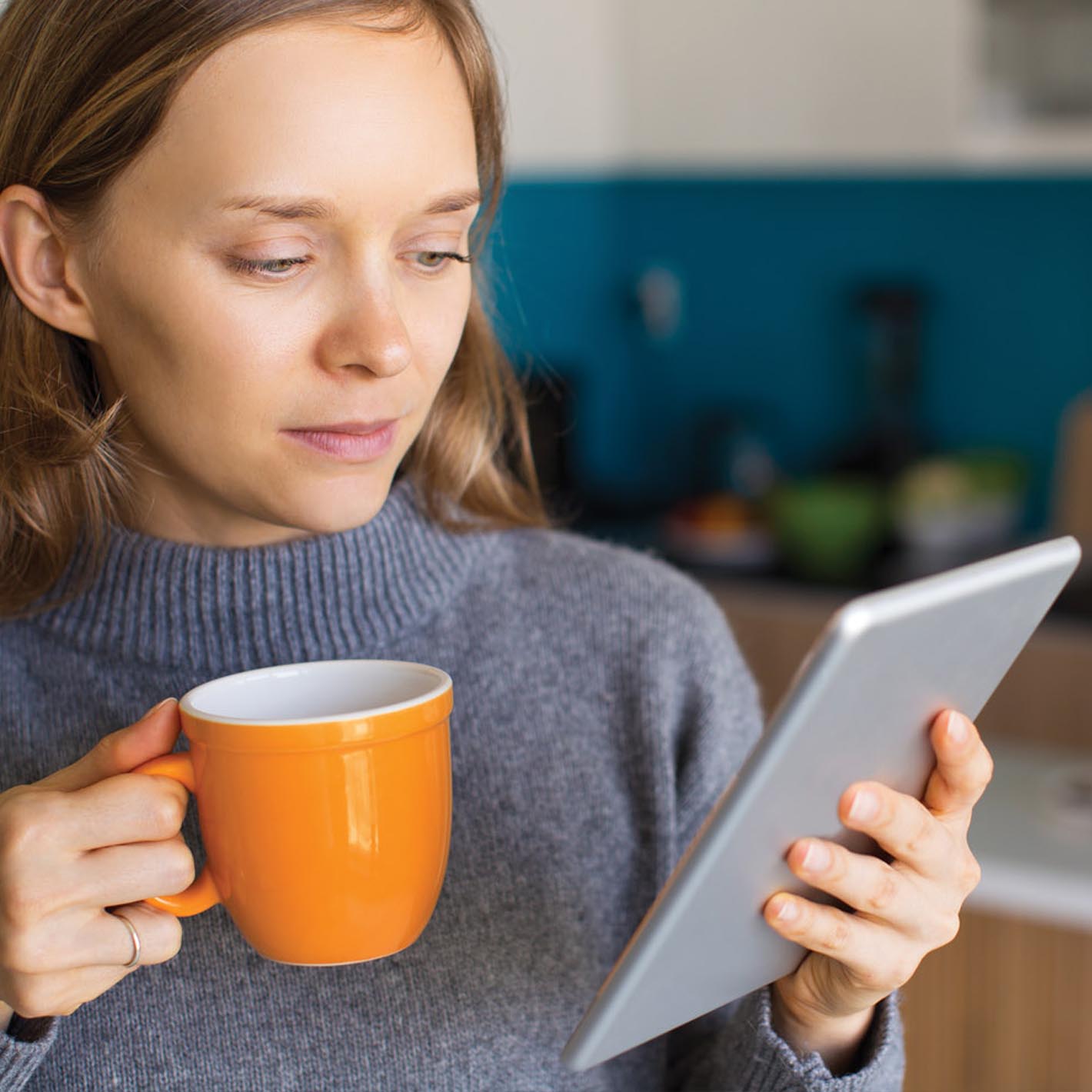 woman holding orange mug and looking at tablet in kitchen